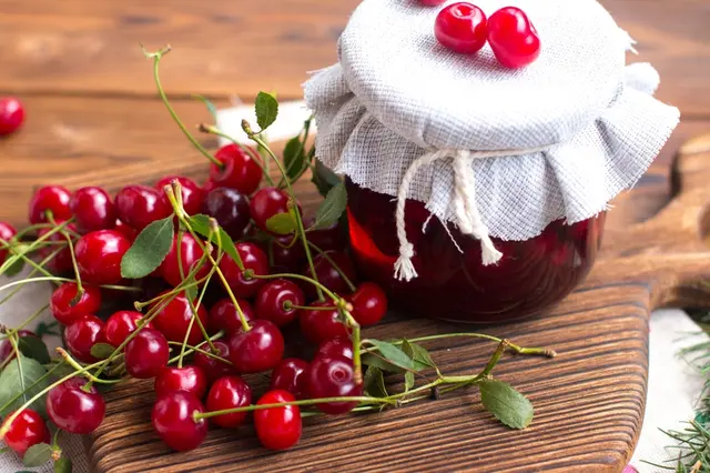 Sklenice s třešňovou marmeládou vedle čerstvých třešní na dřevěném prknu.