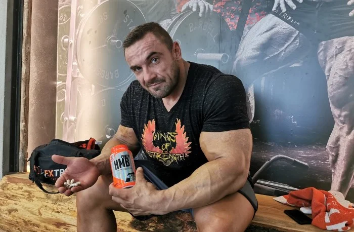 Muž drží nápoj s doplňkem HMB a ukazuje bílé tablety.