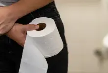 Žena drží záchodový papír a stiská si břicho v koupelně.