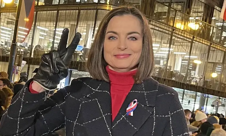 Barbora Kroužková v černém kabátu s červeným svetrem před skleněnou budovou.