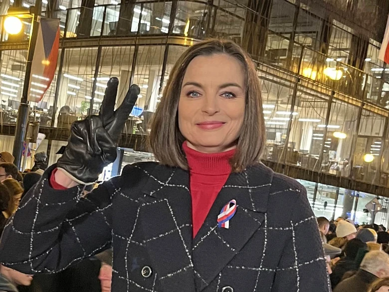 Barbora Kroužková v černém kabátu s červeným svetrem před skleněnou budovou.