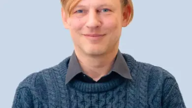 Muž v modrém svetrku se usměvavým výrazem na neutrálním pozadí.