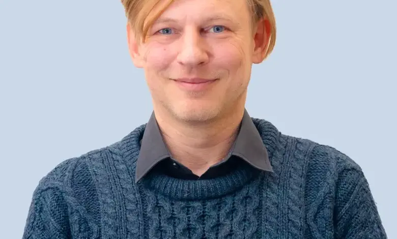Muž v modrém svetrku se usměvavým výrazem na neutrálním pozadí.