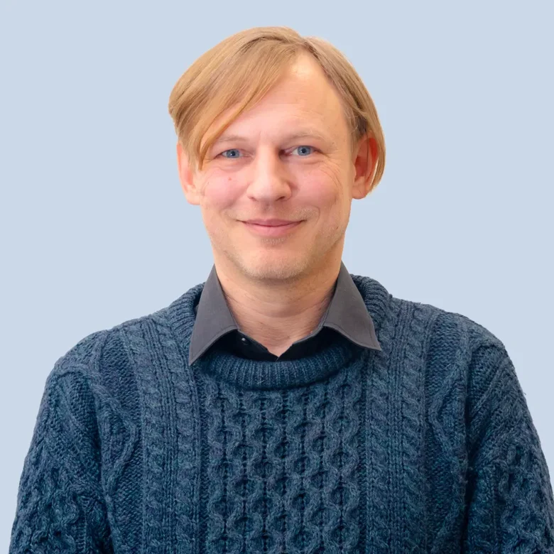 Muž v modrém svetrku se usměvavým výrazem na neutrálním pozadí.